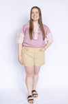 Woman wearing a pink blouse with floral trim and beige shorts 