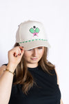 Person wearing a cap with a tennis racket and pink ribbon design 