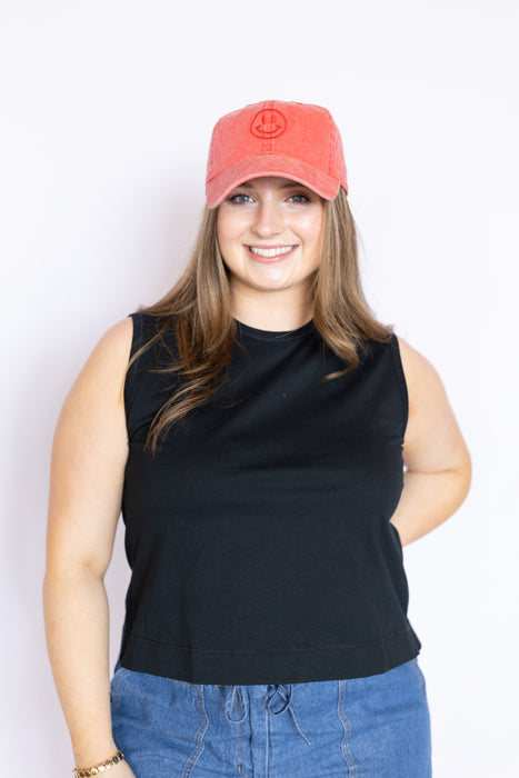 Woman wearing a black sleeveless top and blue jeans with a red smiley face cap 