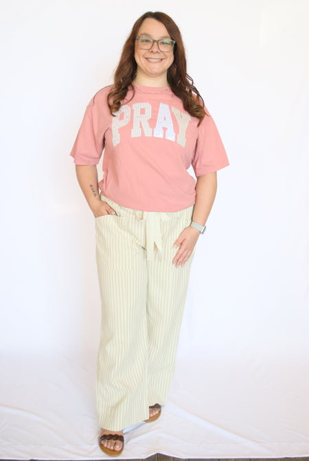 Person wearing a pink 'PRAY' t-shirt and light-colored pants 