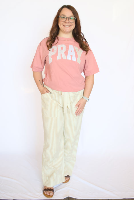 Person wearing a pink 'PRAY' t-shirt and light-colored pants 